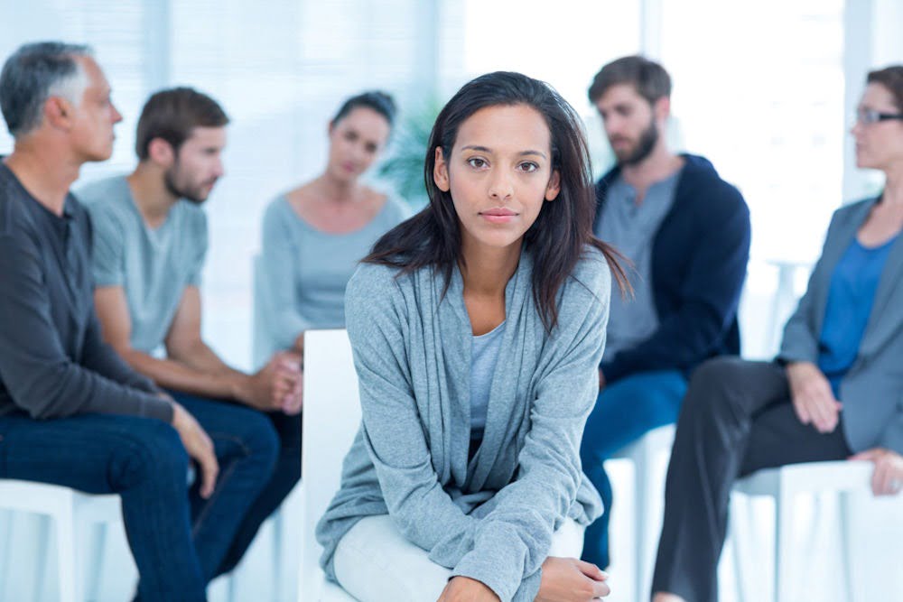 a woman sitting while a group of people on therapy on the background