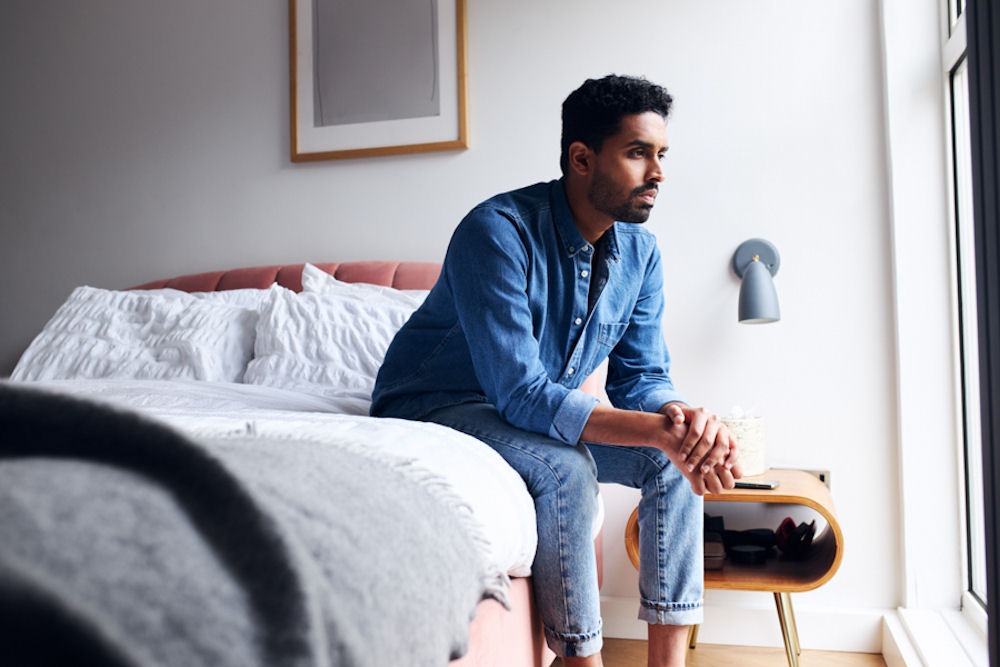 man sitting on the edge of his bed looking toward a window